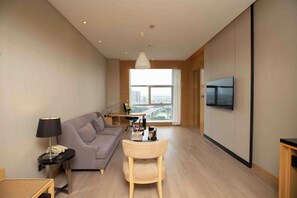 A modern living room with a sofa, a small table, a lamp, a TV mounted on the wall, and a large window offering a city view. - Grand Skylight International Hotel Ganzhou (Ganzhou)