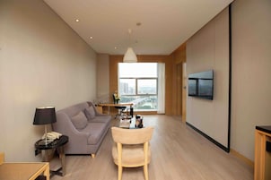 Una sala moderna con sofá, una mesita, una lámpara, televisor montado en la pared y un amplio ventanal con vistas a la ciudad.