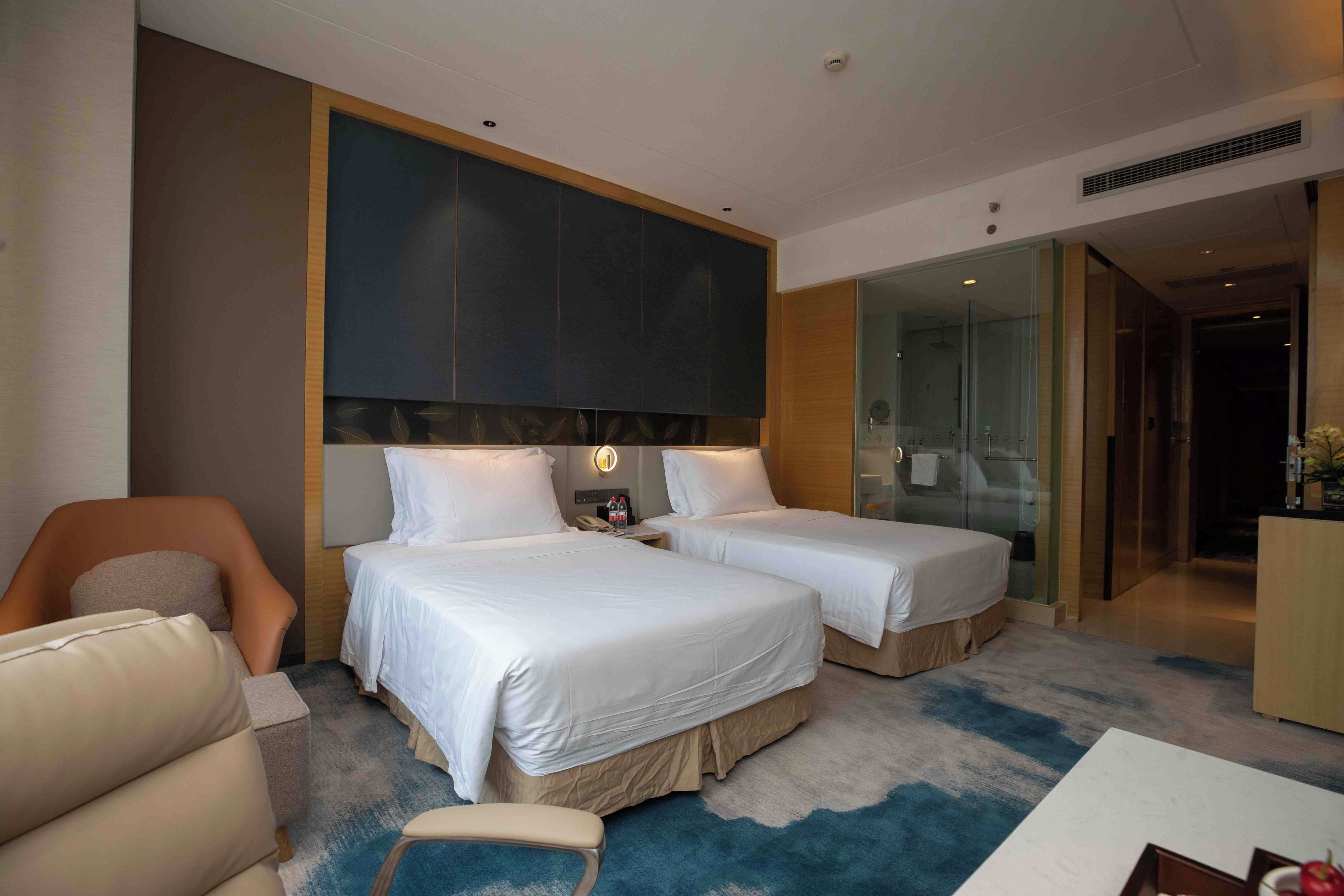 Habitación de hotel con dos camas, un sofá, un escritorio y una zona de ducha con paredes de cristal.