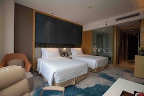 Habitación de hotel con dos camas, un sofá, un escritorio y una zona de ducha con paredes de cristal.