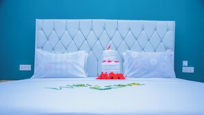 Uma cama com cabeceira estofada, roupa de cama branca e um bolo decorativo como peça central.