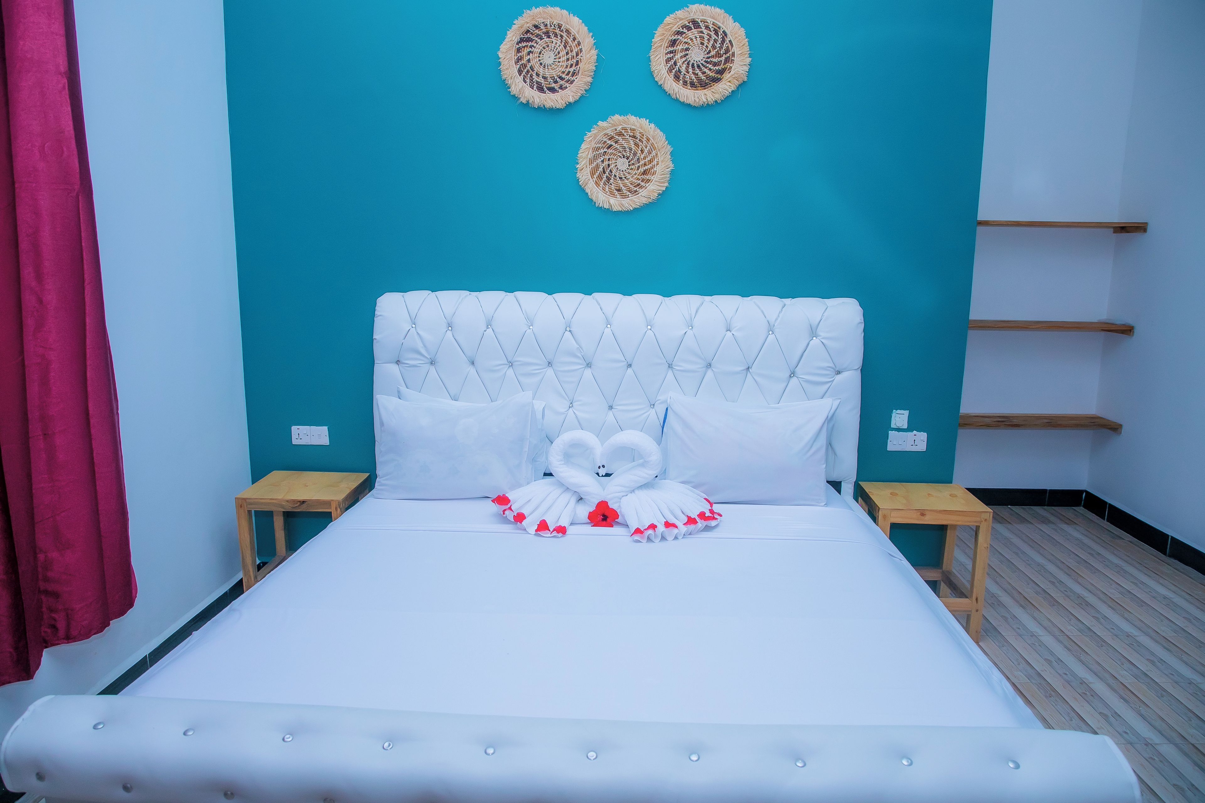 Una cama con cabecera capitonada, ropa de cama blanca y un almohadón con forma de cisne. Hay dos mesitas de noche, una pared color turquesa y una cortina roja.