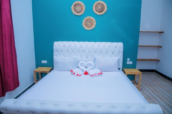 A bed with a tufted headboard, white bedding, and a swan-shaped pillow. There are two bedside tables, a teal wall, and a red curtain. - Mango Grove Villa (Uroa)