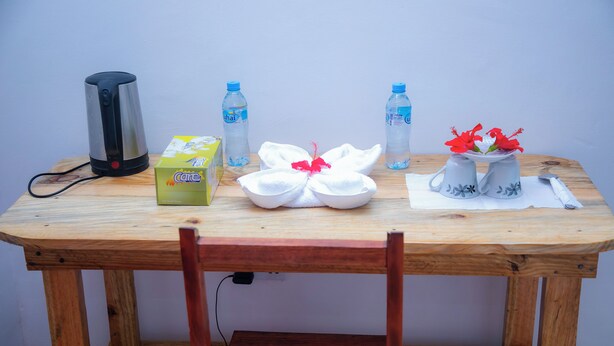 Uma mesa de madeira com um porta-lenços, duas garrafas de água, uma chaleira, uma tigela com uma flor vermelha e duas canecas com estampas florais.