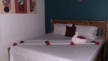 Cama bem arrumada com lençóis brancos e flores vermelhas, cabeceira de madeira, e parede com arte abstrata e tapeçarias.