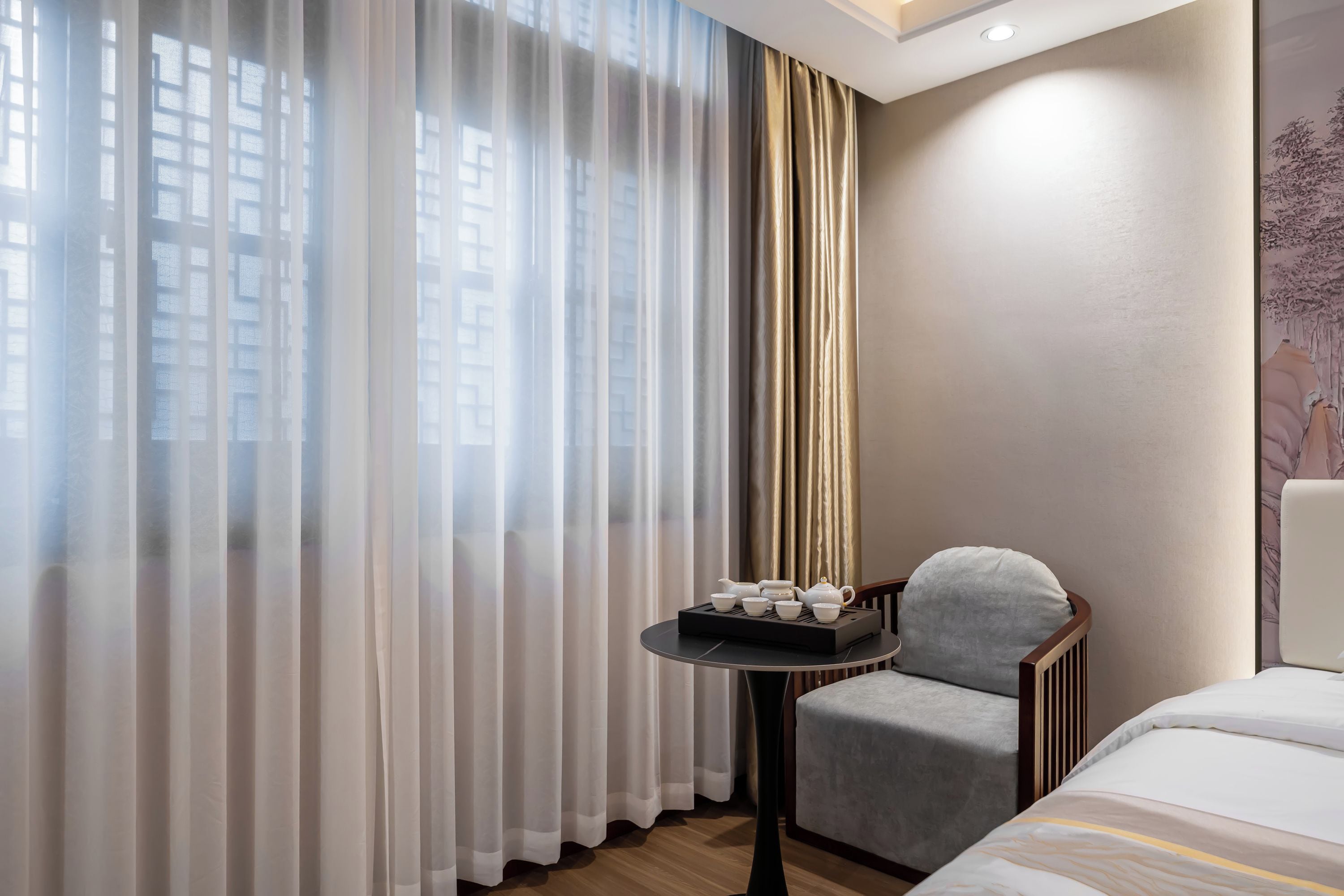 A hotel room with a bed, a chair, a small table with tea set, and a view through sheer curtains.