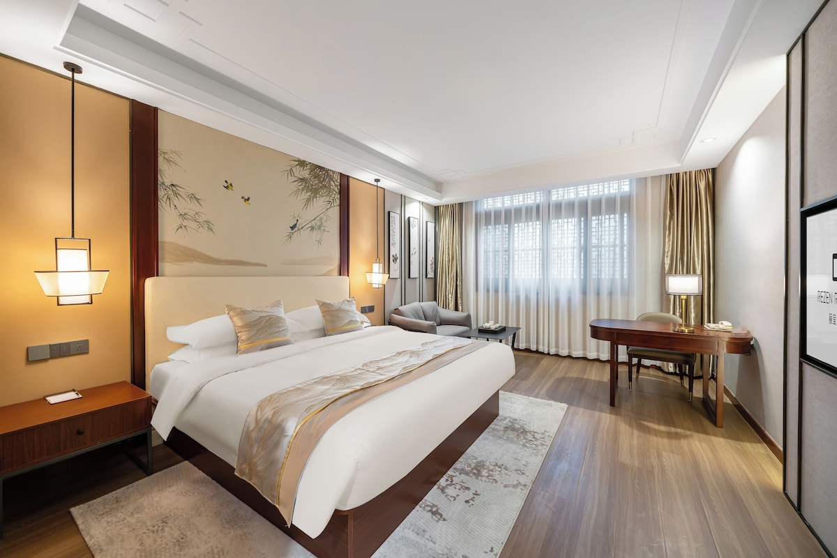 Quarto de hotel com uma cama grande, mesas de cabeceira, escrivaninha e vista para a cidade pela janela.