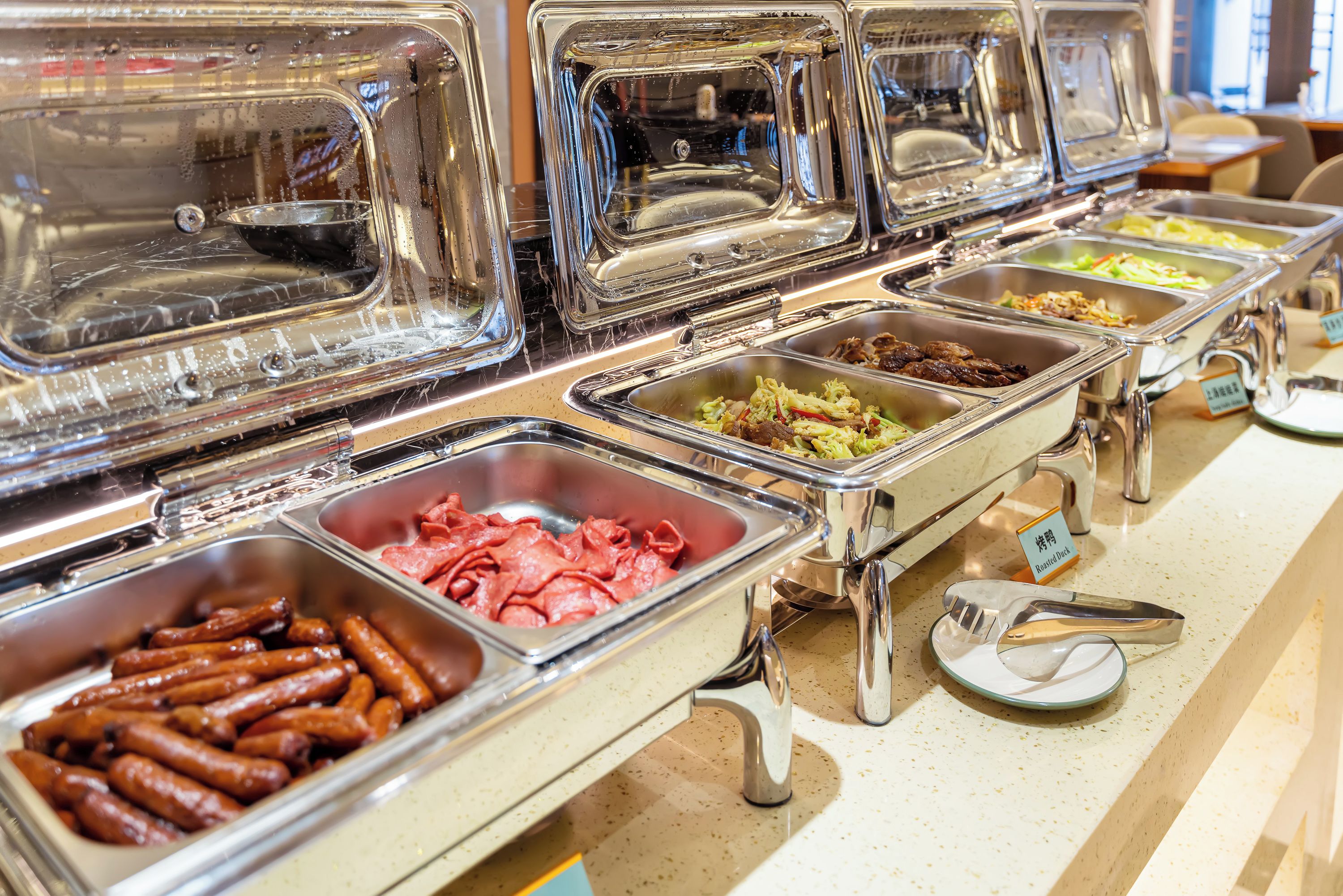 Un bufé con diversas opciones de comida, incluyendo salchichas, pato asado y verduras.