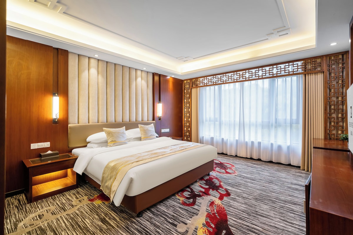 Quarto de hotel com uma cama grande, mesa de cabeceira, escrivaninha e janela com cortinas.