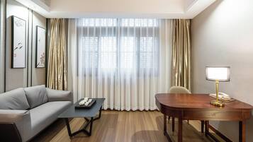 A hotel room with a sofa, a small table, a desk with a lamp, and a window with curtains.