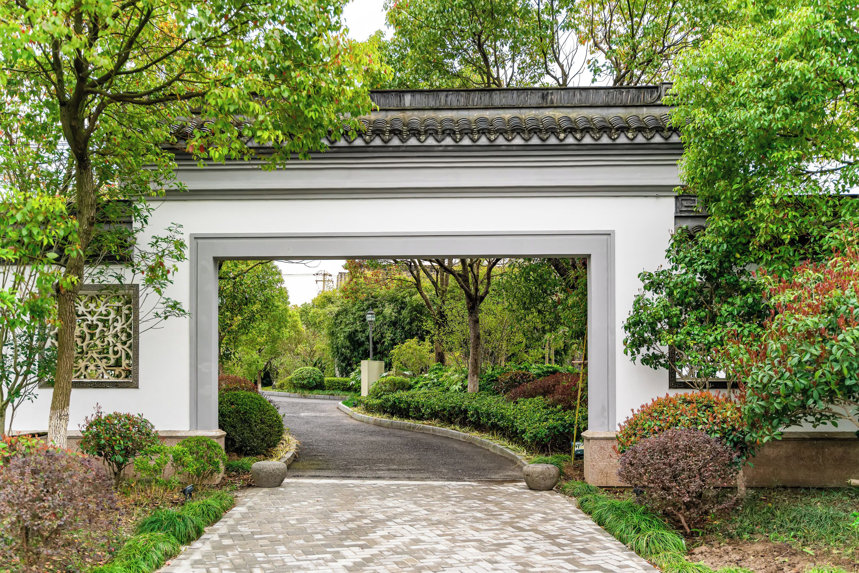 A traditional gate with a tiled roof, surrounded by lush greenery and a paved pathway leading through it.