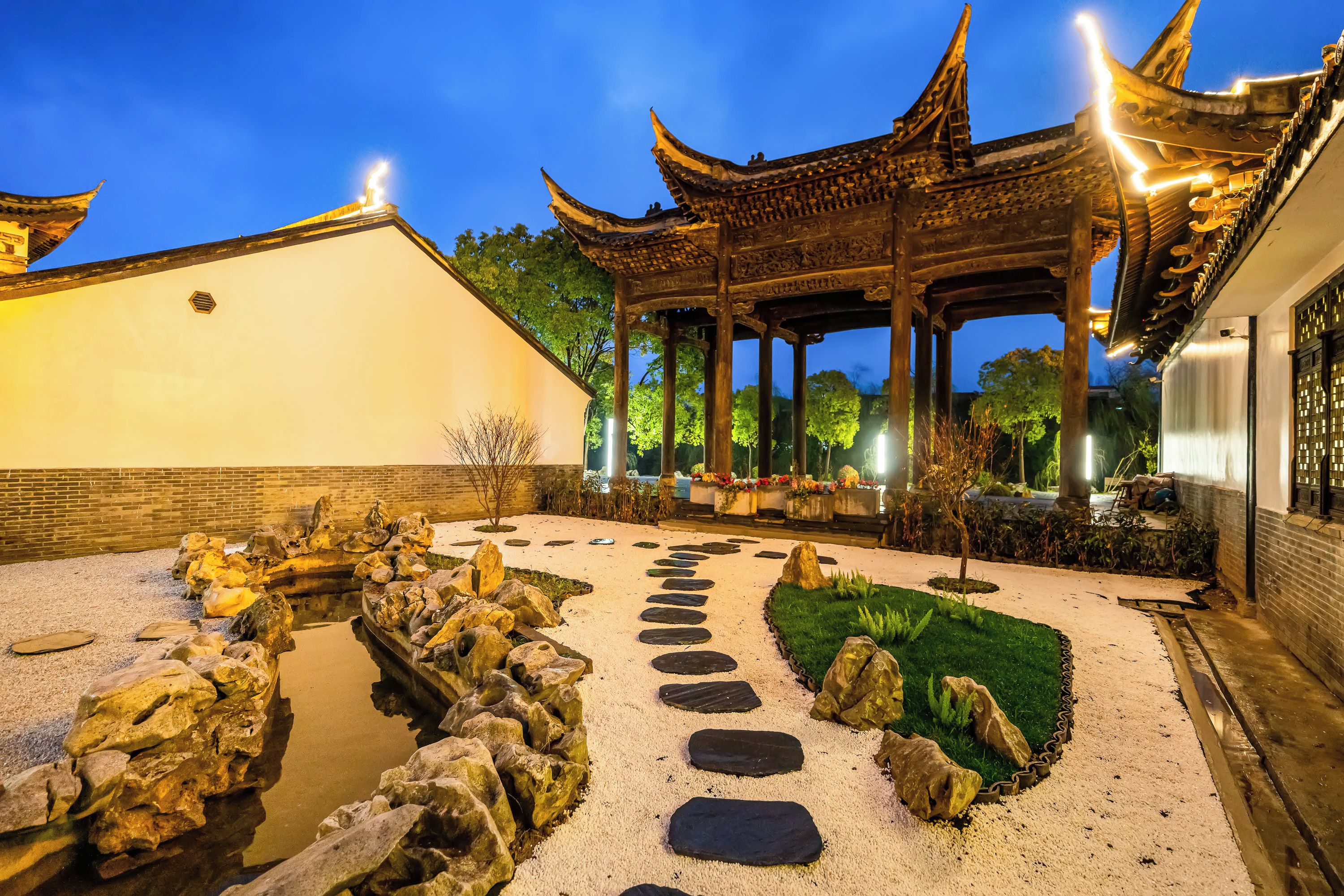 A traditional pavilion with a tiled roof, surrounded by a gravel path with stepping stones, and a small water feature with rocks.