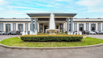 A building with a central fountain, surrounded by a manicured lawn and a parking lot with several cars.