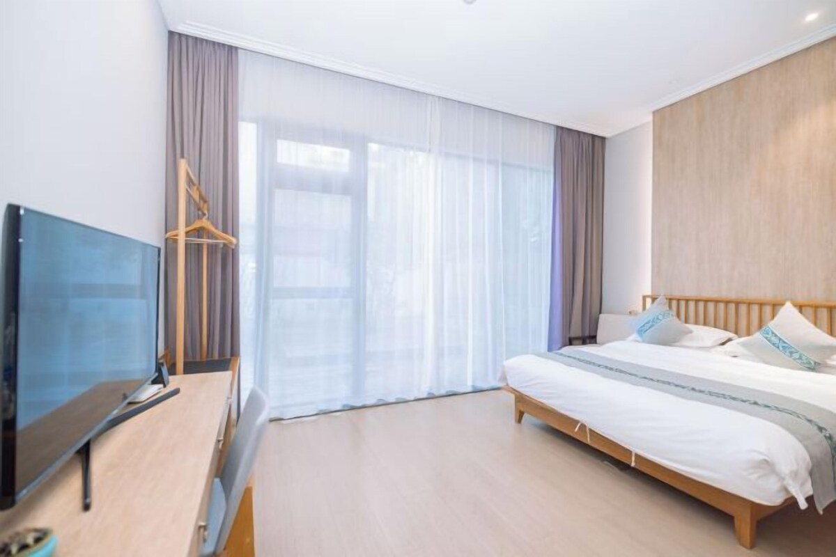 Habitació d'hotel moderna amb un llit, una televisió de pantalla plana, un escriptori de fusta i una finestra gran amb cortines transparents.