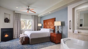 Habitación de hotel con cama, chimenea, ventana grande con cortinas, ventilador de techo, mesita de noche, lámpara, bañera y espejo.