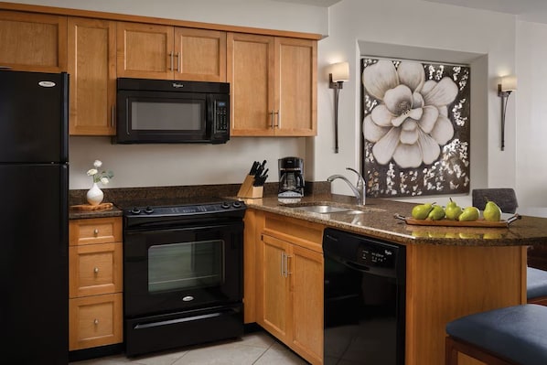 Cocina con armarios de madera, refrigerador negro, microondas, horno, lavavajillas y encimera de granito con un frutero.