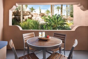 Una mesa redonda de madera con cuatro sillas, una pequeña bandeja de madera con dos vasos y una flor roja, y una vista de un paisaje rocoso con palmeras a través de una ventana.