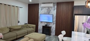 A living room with a sofa, ottoman, TV, fireplace, and a dining area with a table and chairs.