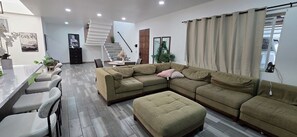 A modern living room with a sectional sofa, a dining area with chairs, a staircase, and a large window with curtains.