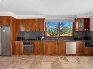 Cocina moderna con armarios de madera, un refrigerador de acero inoxidable, horno empotrado, lavavajillas y un amplio ventanal con vista a árboles.