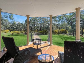 Un espacio de estar al aire libre cubierto con mobiliario de mimbre y una mesa de vidrio, con vistas a un jardín cuidado y un sendero.