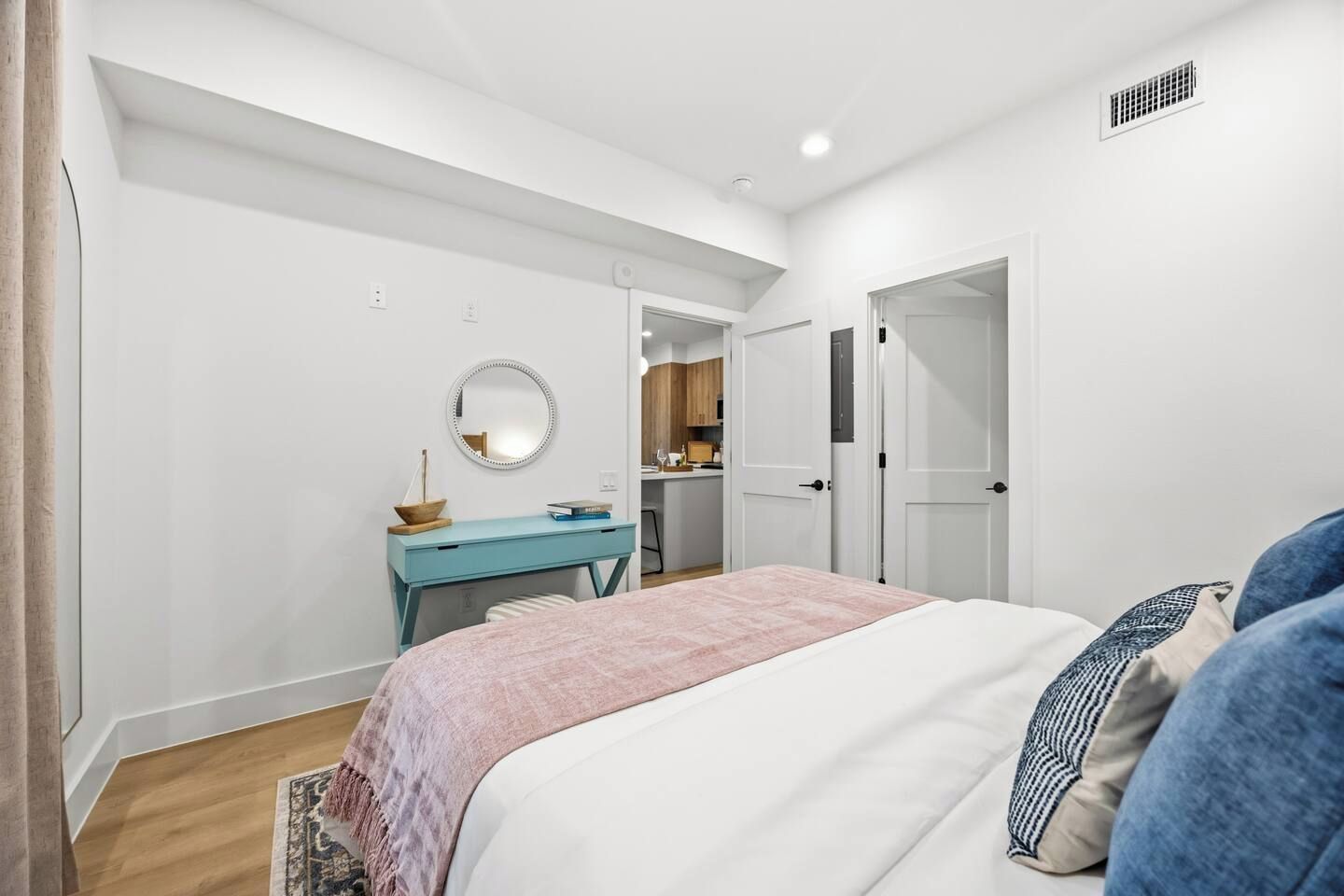 A bedroom with a bed, a desk, a mirror, and a door leading to a kitchen.
