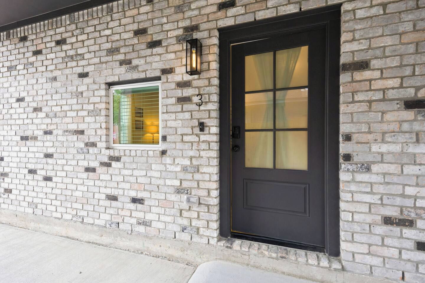 Un bâtiment en briques avec une porte noire et une fenêtre dotée d’un luminaire jaune.