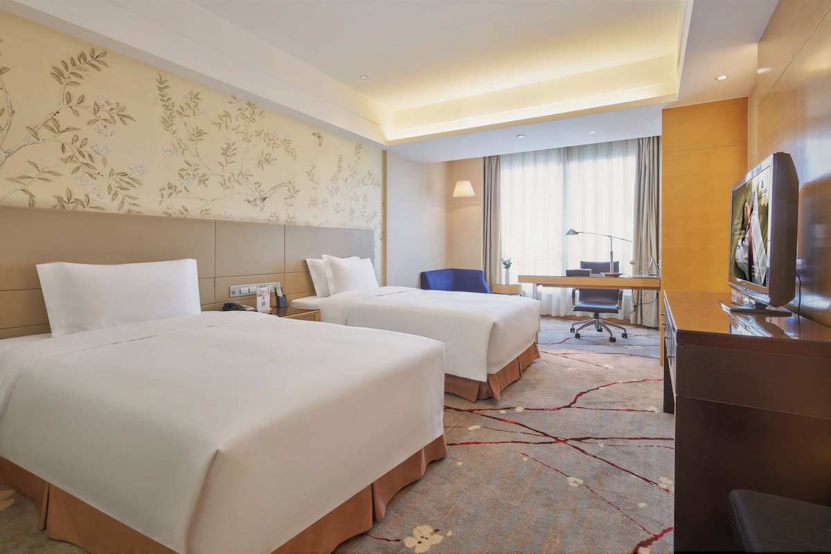 Habitación de hotel con dos camas, un escritorio, una silla, un televisor y un ventanal con cortinas.