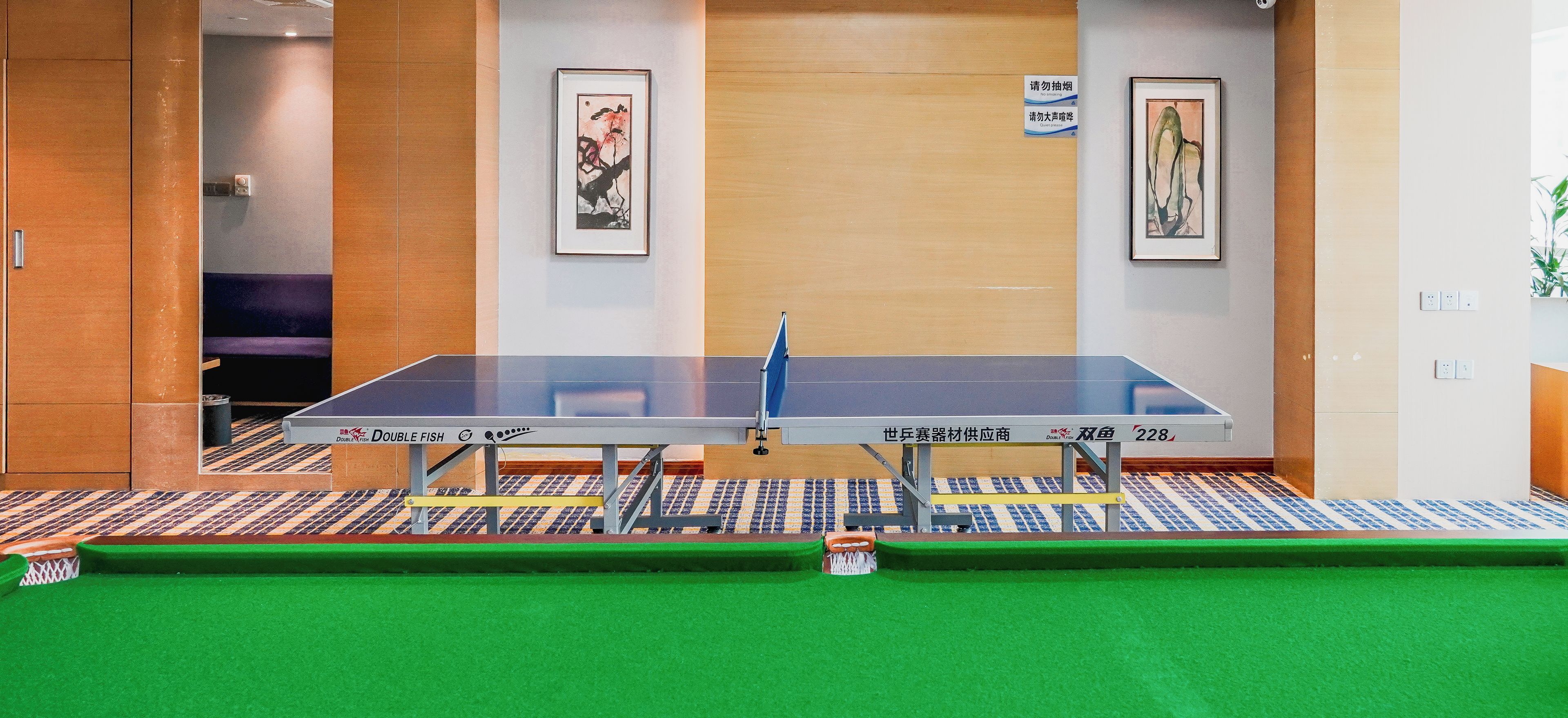 Une table de ping-pong dans une pièce dont le sol est recouvert d’une moquette verte, deux œuvres d’art encadrées accrochées au mur et un panneau indiquant qu’il est interdit de fumer.