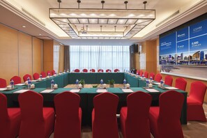 A conference room with a long table, red chairs, and a large screen displaying hotel information.