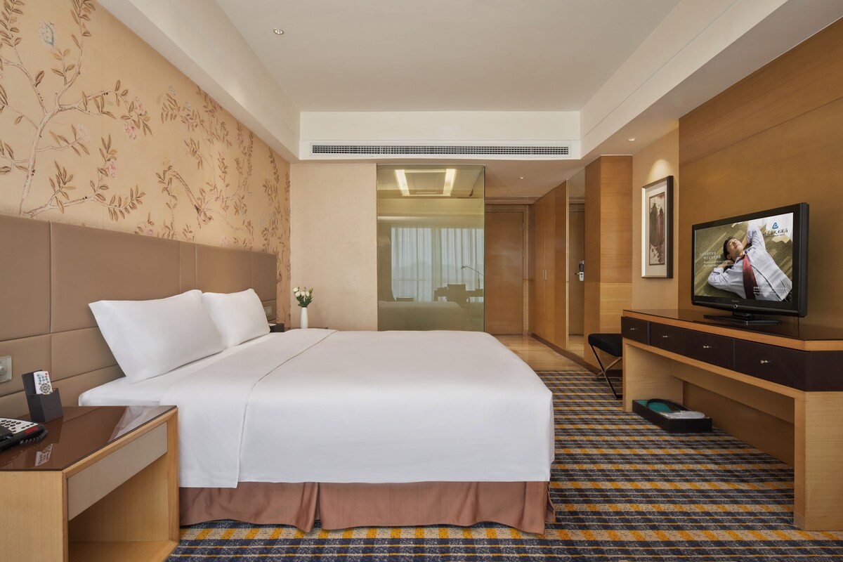 Habitación de hotel con una cama grande, televisión, escritorio y baño visible a través de una puerta de cristal.