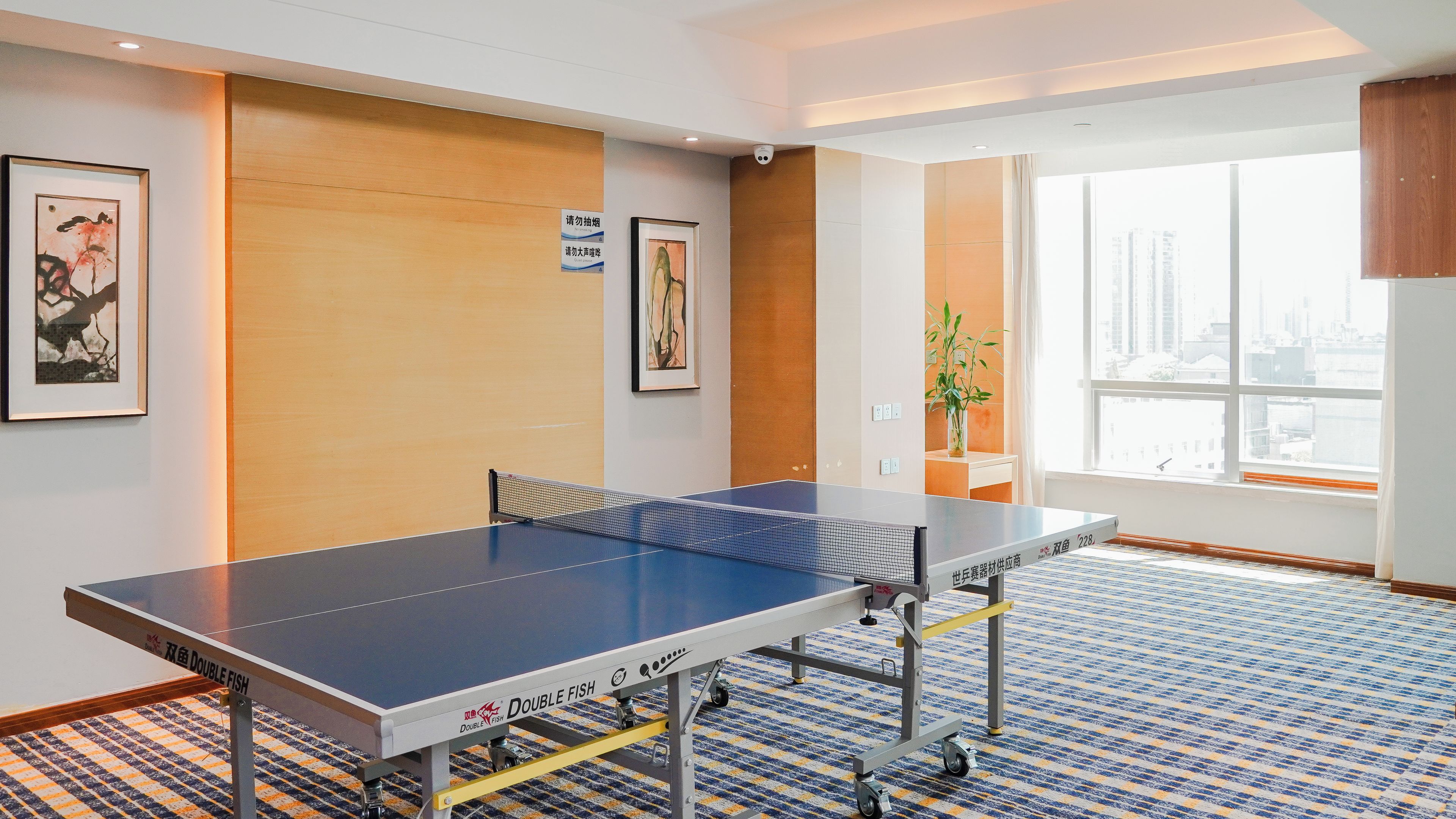 A ping pong table in a hotel room with a city view, a framed artwork on the wall, and a potted plant by the window.