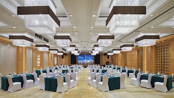 Un salón de conferencias con filas de mesas y sillas, candelabros y una pantalla grande al frente.