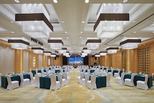 A conference room with rows of tables and chairs, chandeliers, and a large screen at the front.