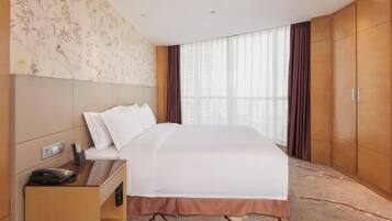 Quarto de hotel com uma cama grande, mesa de cabeceira com telefone e controle remoto, TV na parede e janela com cortinas.
