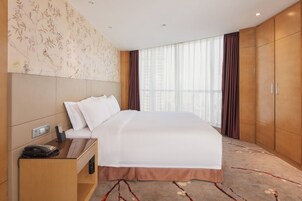 Quarto de hotel com uma cama grande, mesa de cabeceira com telefone e controle remoto, TV na parede e janela com cortinas.