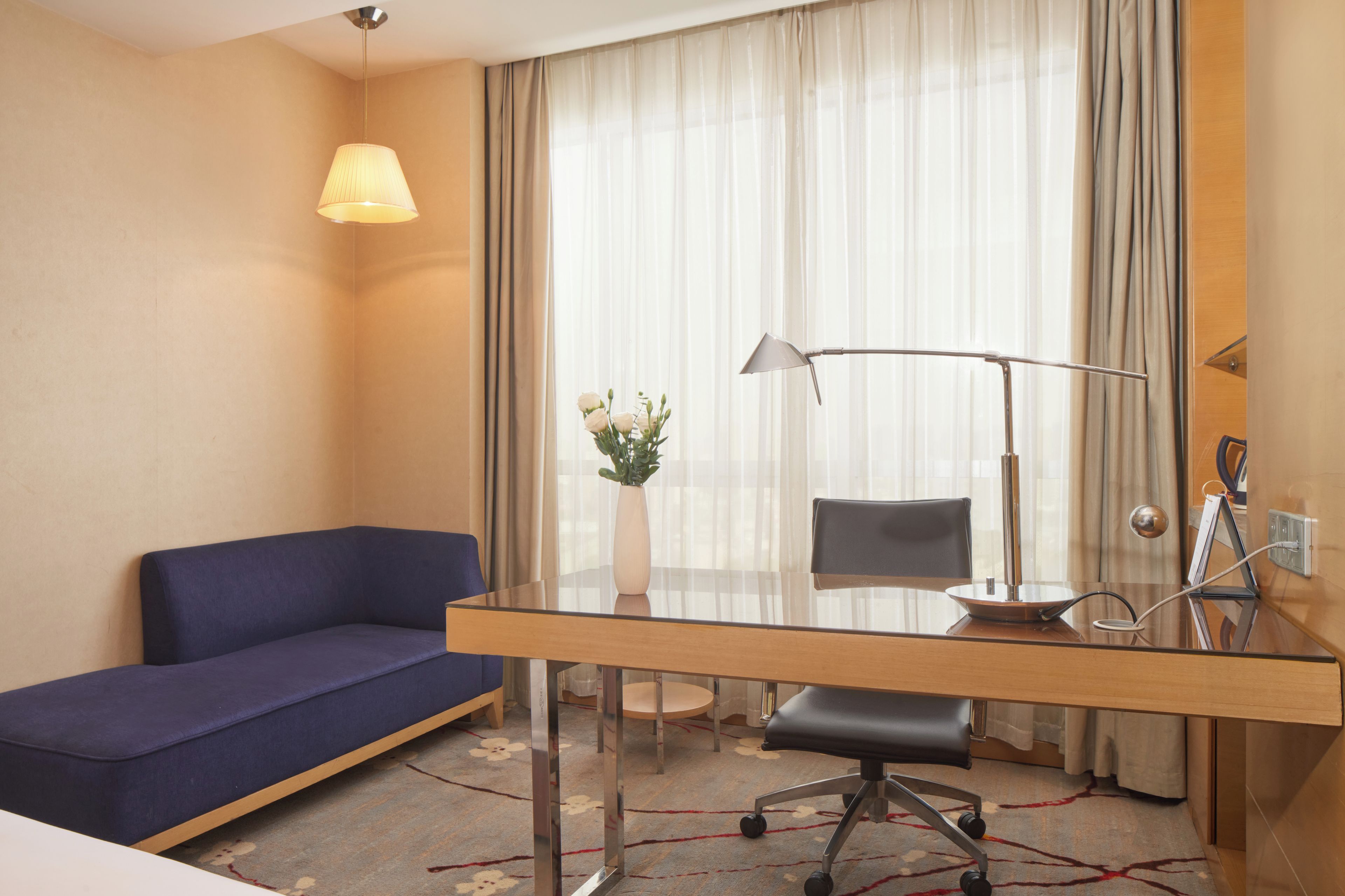 A hotel room with a sofa, a desk with a lamp, a chair, a vase with flowers, and a window with curtains.