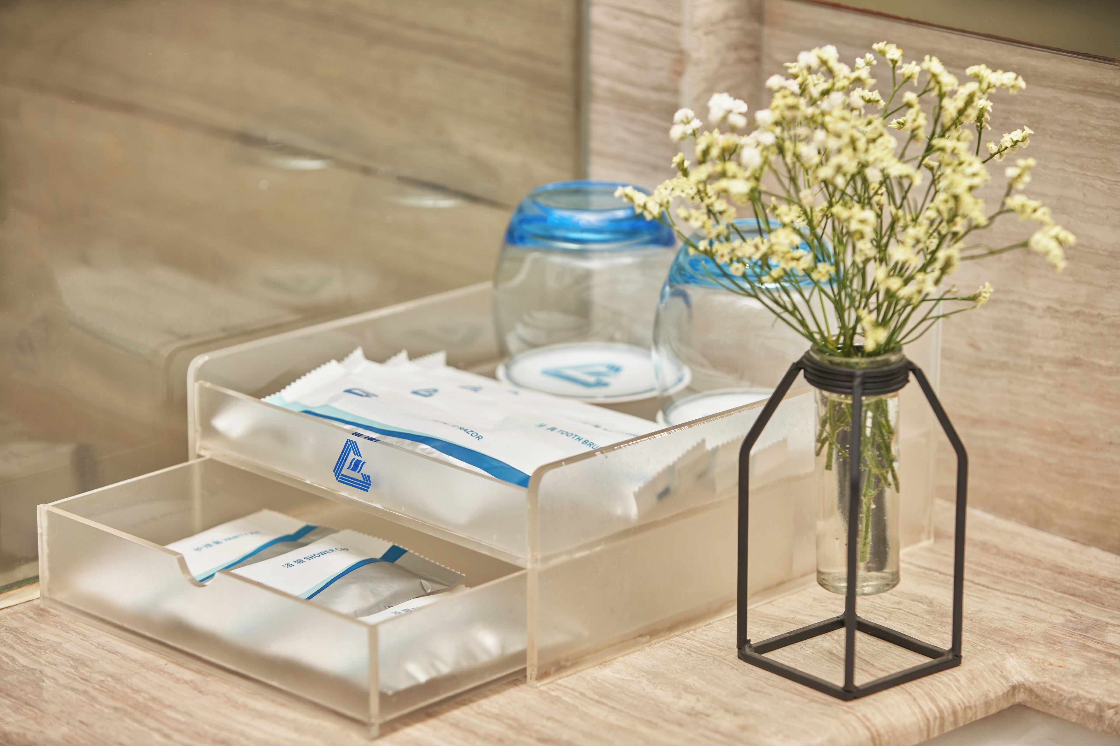 En genomskinlig plastförvaringslåda med fack som innehåller toalettartiklar, en glasburk och en vas med blommor.