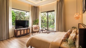 Quarto de hotel com uma cama grande, televisão, área de estar com cadeira e mesa, e varanda com vista para vegetação.