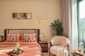 A hotel room with a bed, a chair, a bedside table, a lamp, a plant, and a window with curtains.