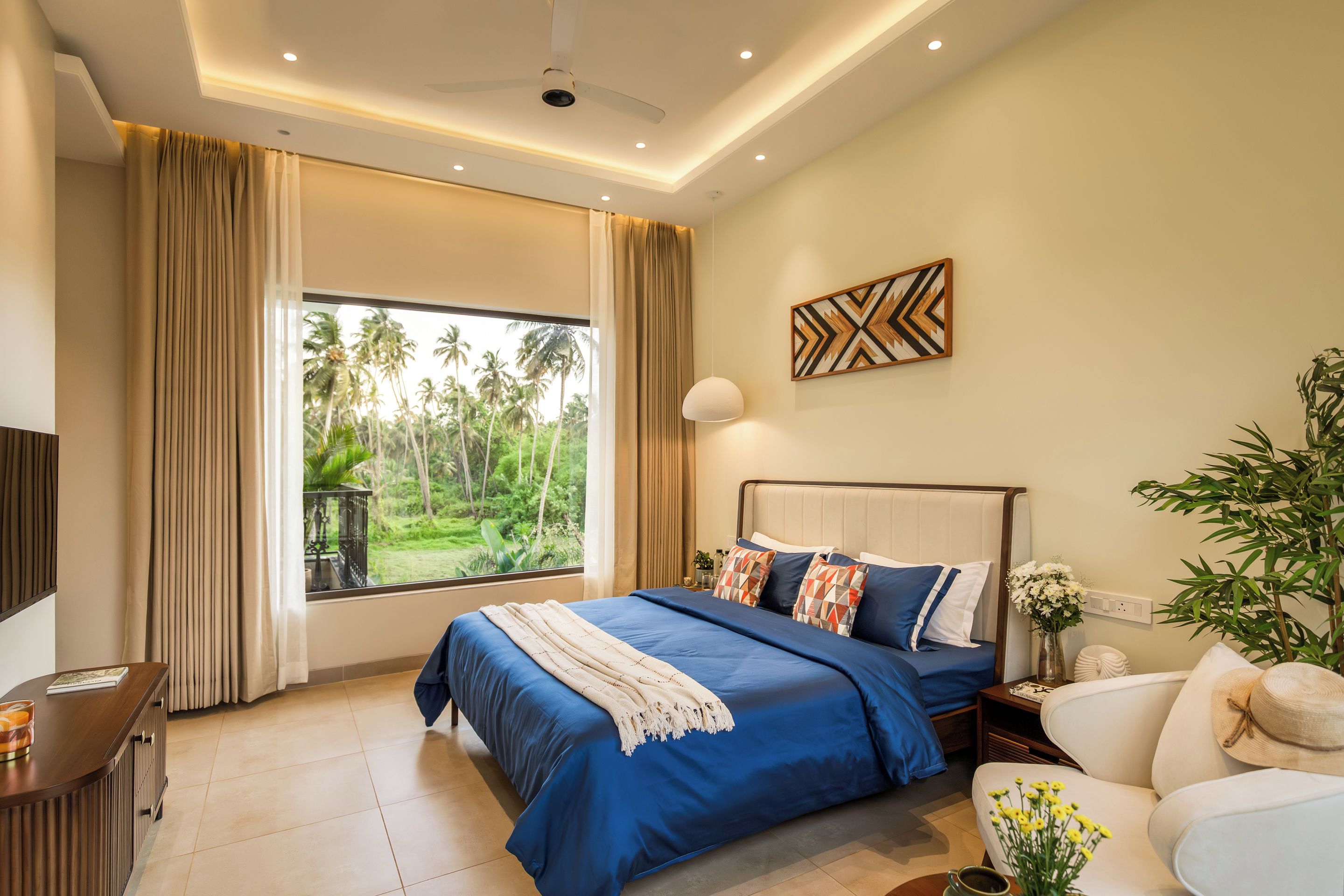 Quarto com uma grande janela, cama com roupa de cama azul, mesa de cabeceira, abajur e vista para palmeiras.