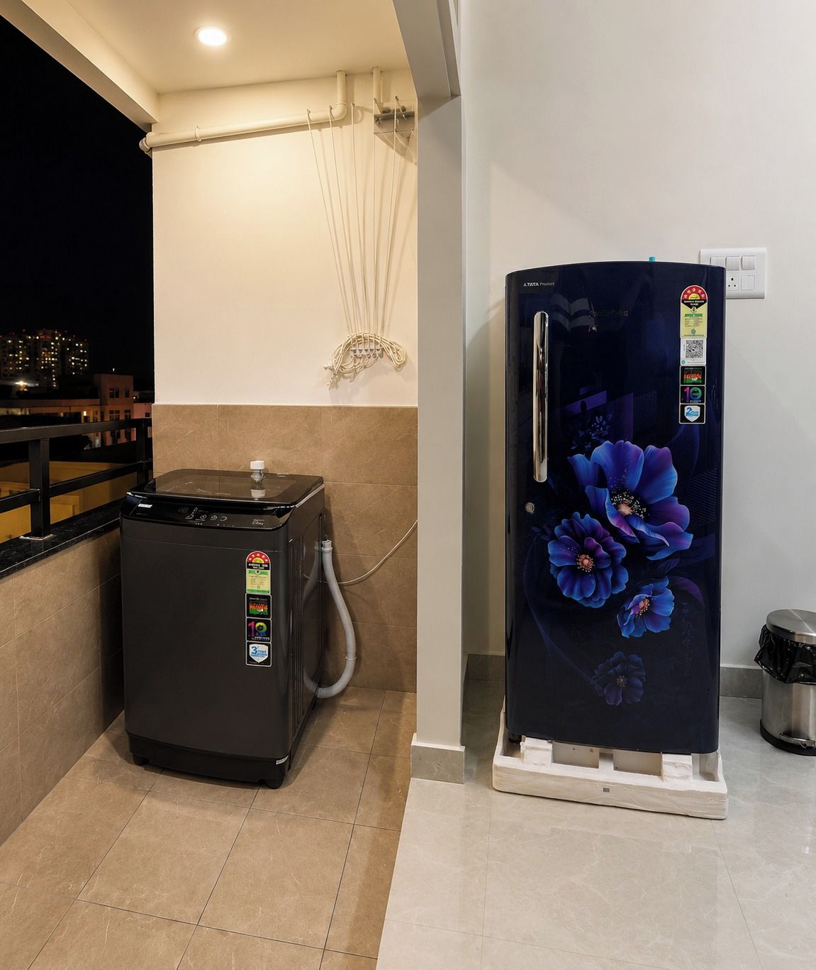 Una cocina con un cocedor de arroz negro, un refrigerador con diseño floral y una tubería de agua visible en la pared.