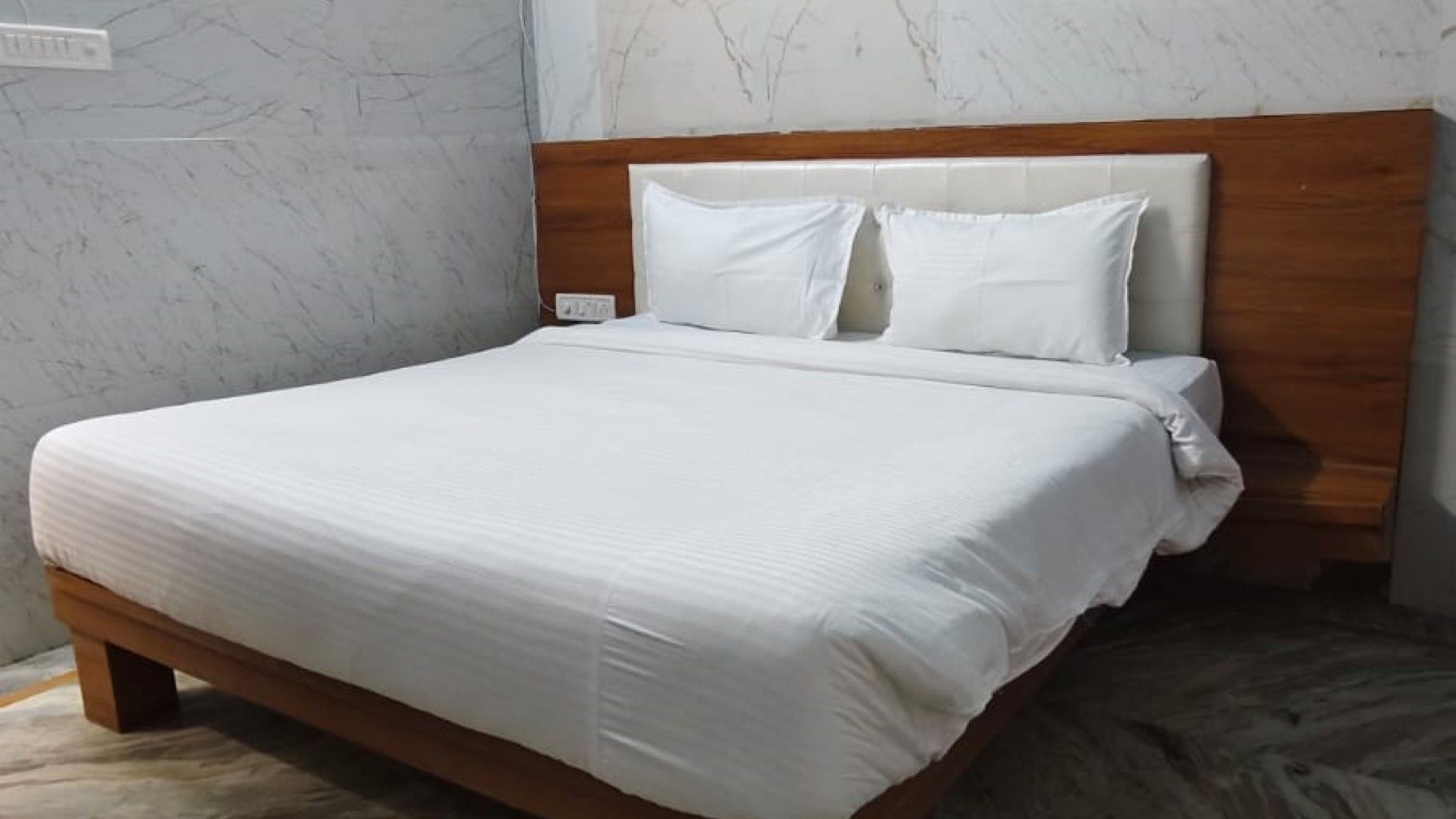 Una cama bien tendida con cabecera de madera, ropa blanca y una pared con diseño de mármol.
