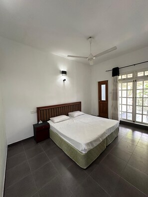 Une chambre à coucher avec un lit, une table de chevet, un ventilateur de plafond et une fenêtre avec des rideaux.