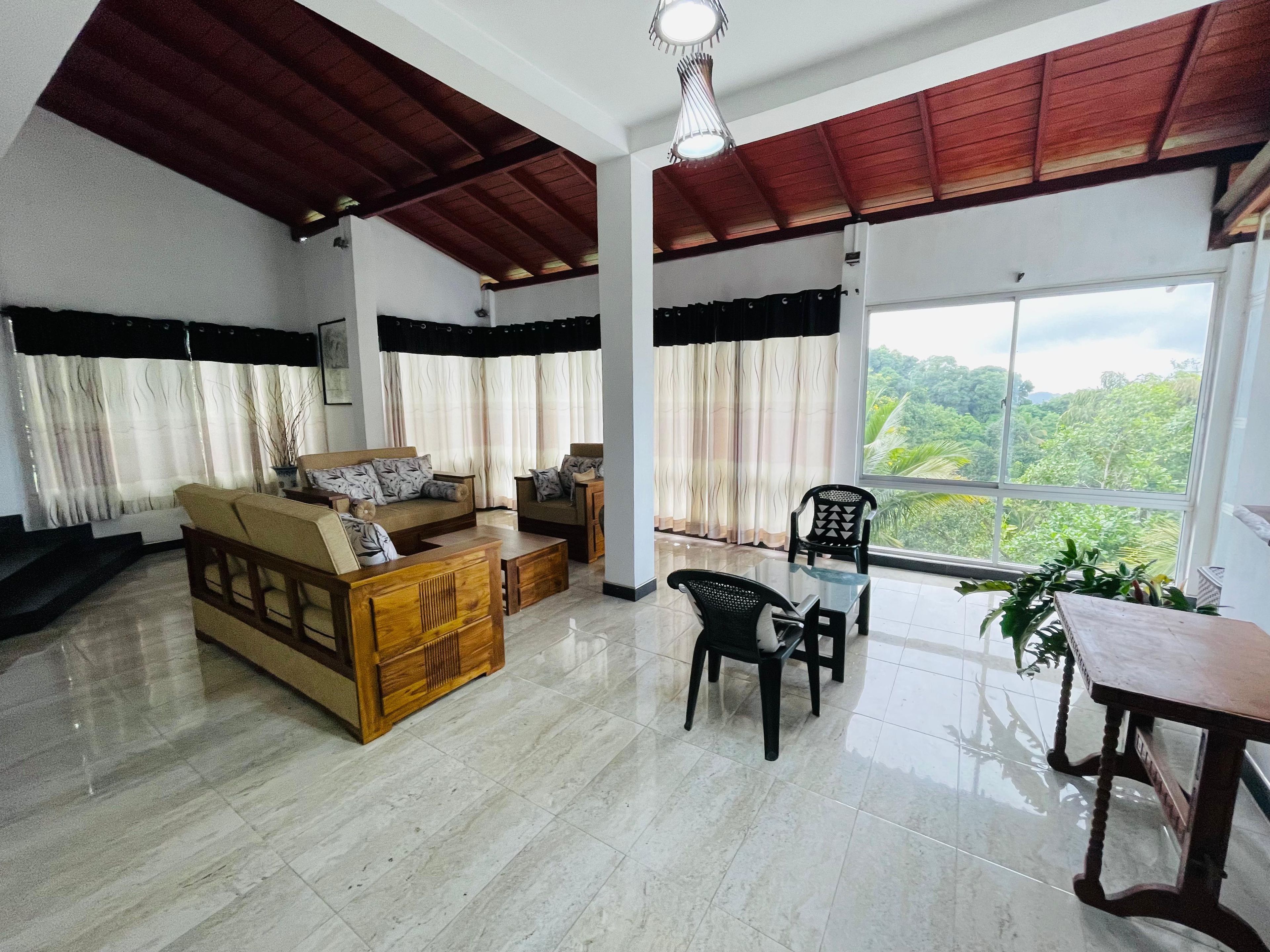 A spacious living area with a sofa set, a coffee table, and a dining table with chairs. There is a large window offering a view of greenery outside.