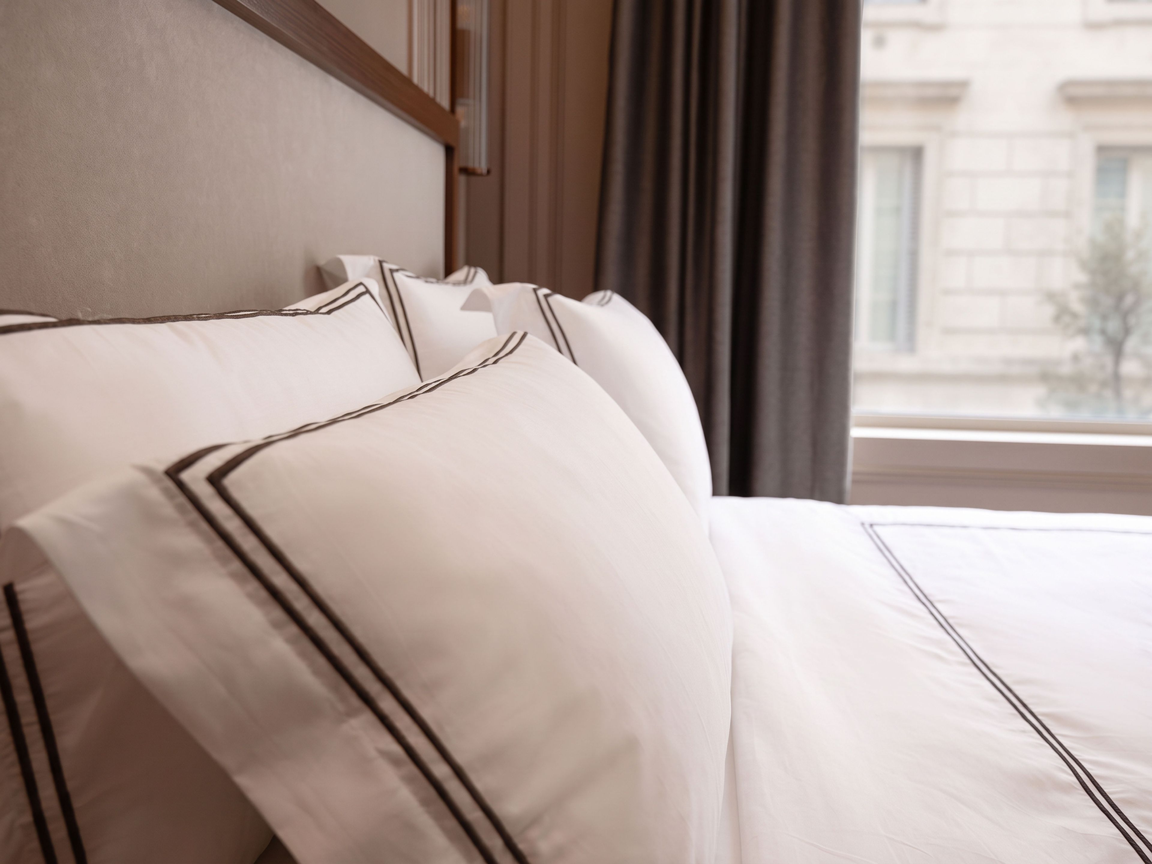 A neatly made bed with white linens and dark trim, a wooden headboard, and a window with curtains.