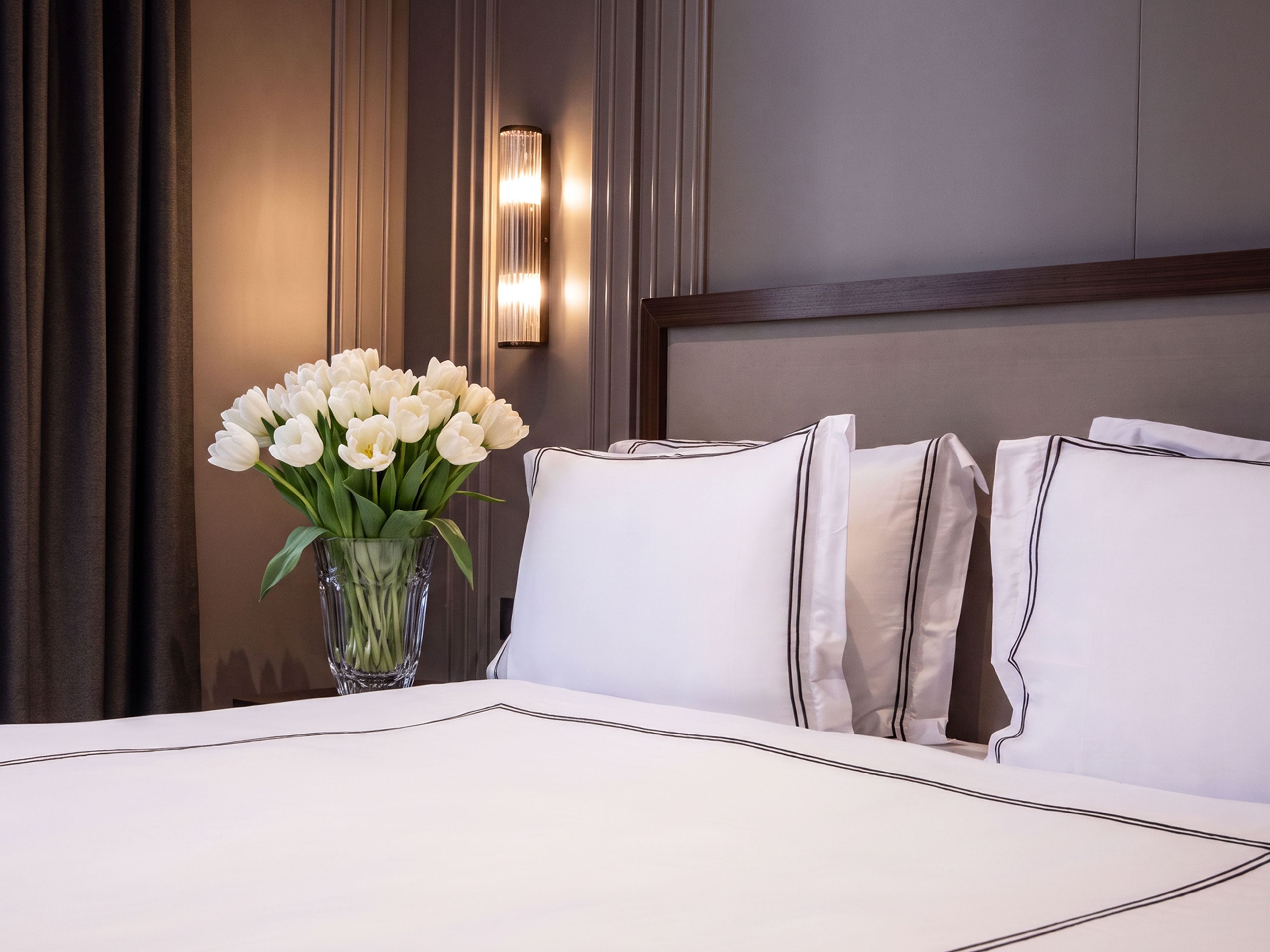 Une chambre d’hôtel avec un lit, des oreillers blancs, un vase avec des fleurs blanches et un luminaire fixé au mur.
