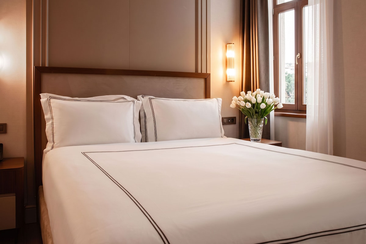Une chambre d’hôtel avec un grand lit, une table de chevet, un vase de fleurs et une fenêtre avec des rideaux.