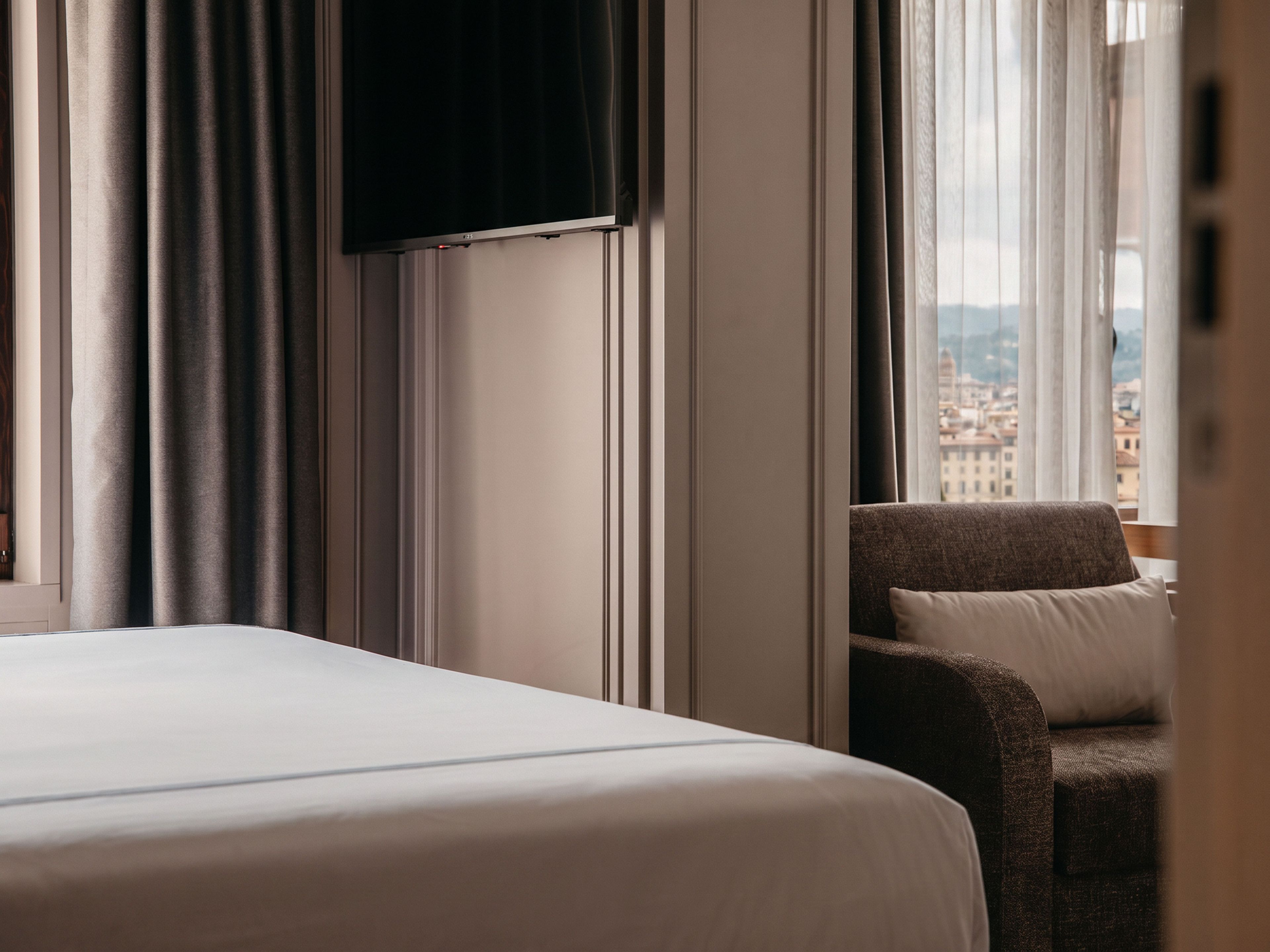 A hotel room with a bed, a chair, a television, and a view of a cityscape through the window.