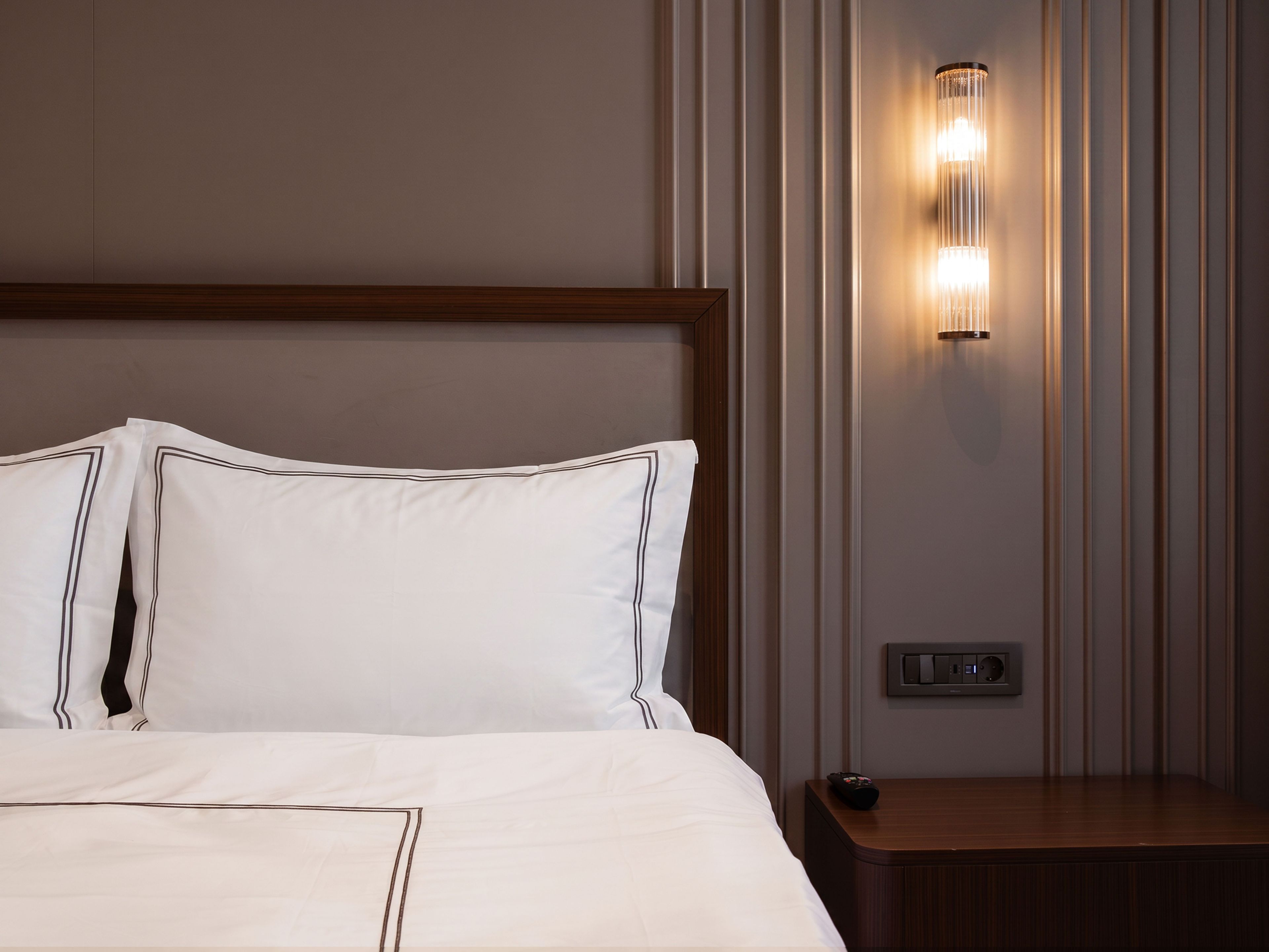 A hotel room with a neatly made bed, a bedside table, and a wall-mounted light fixture.